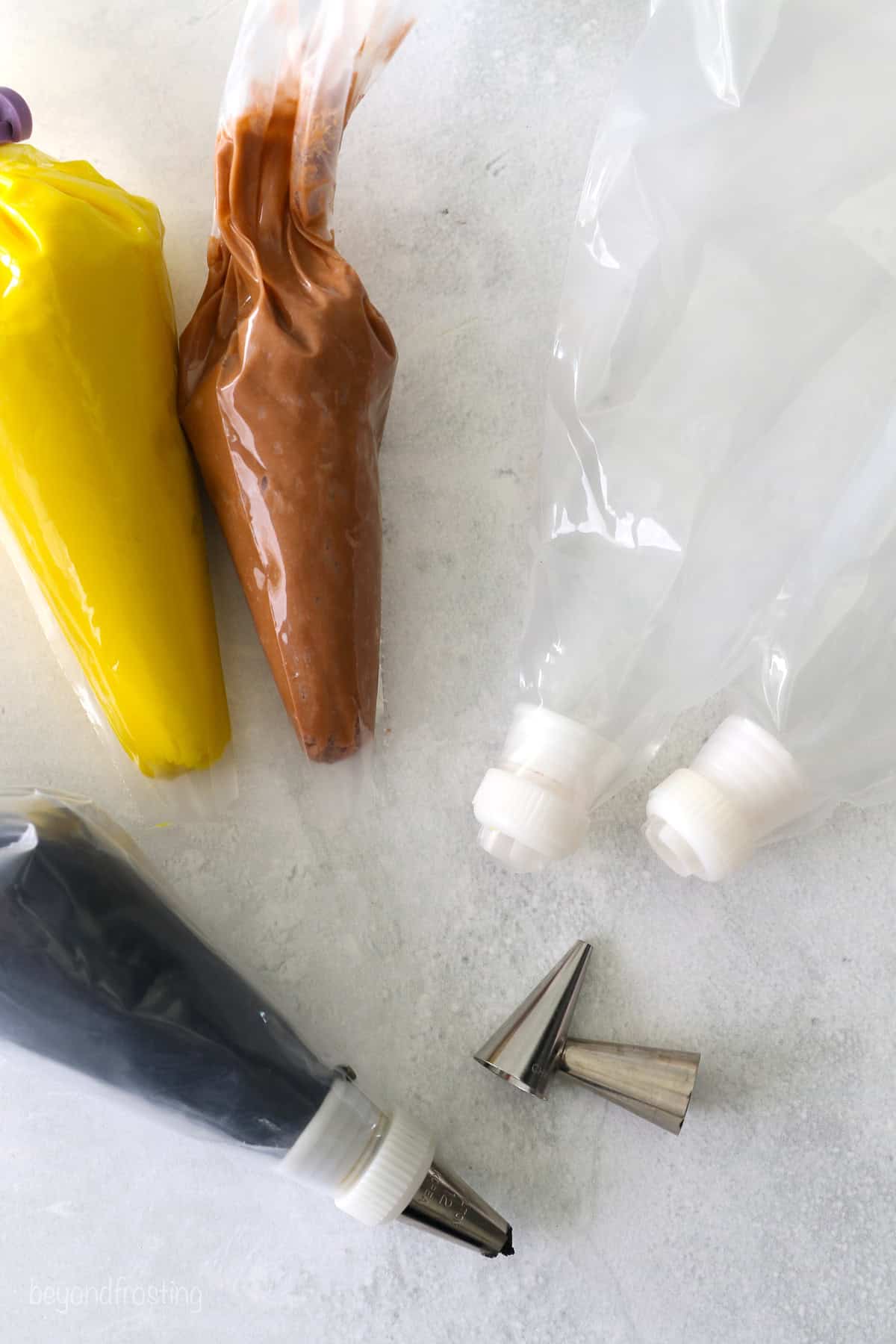 Overhead view of three piping bags filled with yellow, brown, and black buttercream frosting next to two empty piping bags fitted with couplers and two scattered piping tips.