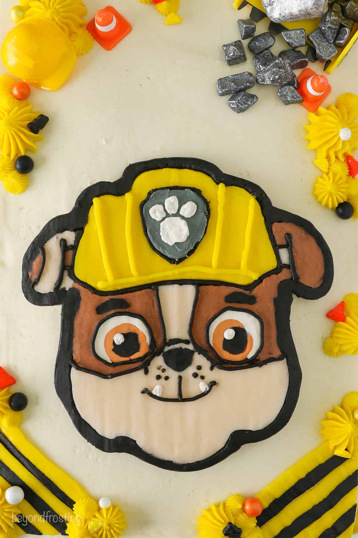 Overhead view of a Rubble Paw Patrol Cake with Rubble's detailed face in the center, surrounded by piped buttercream shells and construction decorations.