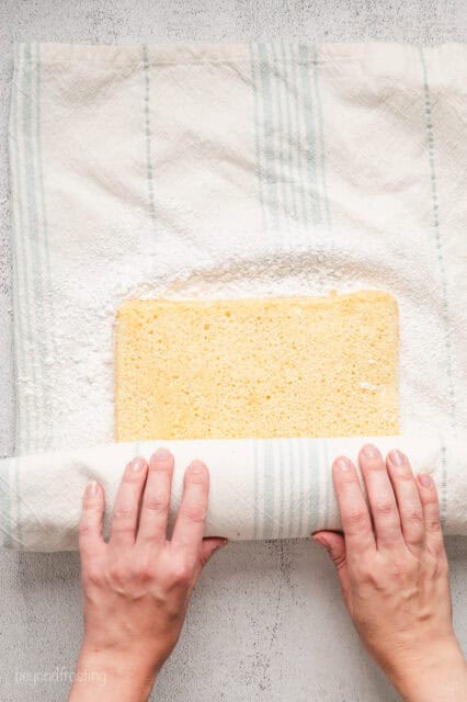 Two hands rolling a sheet of vanilla cake inside a dish towel.