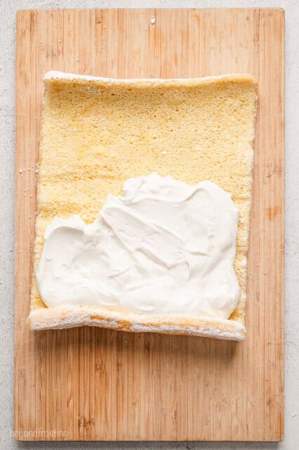 Overhead view of a pre-rolled vanilla cake sheet partially frosted with cream cheese frosting.
