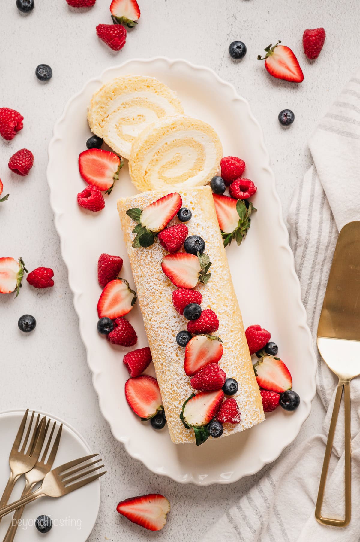 Overhead view of a vanilla cake roll topped with fresh berries, with two slices cut from the end on a large platter.