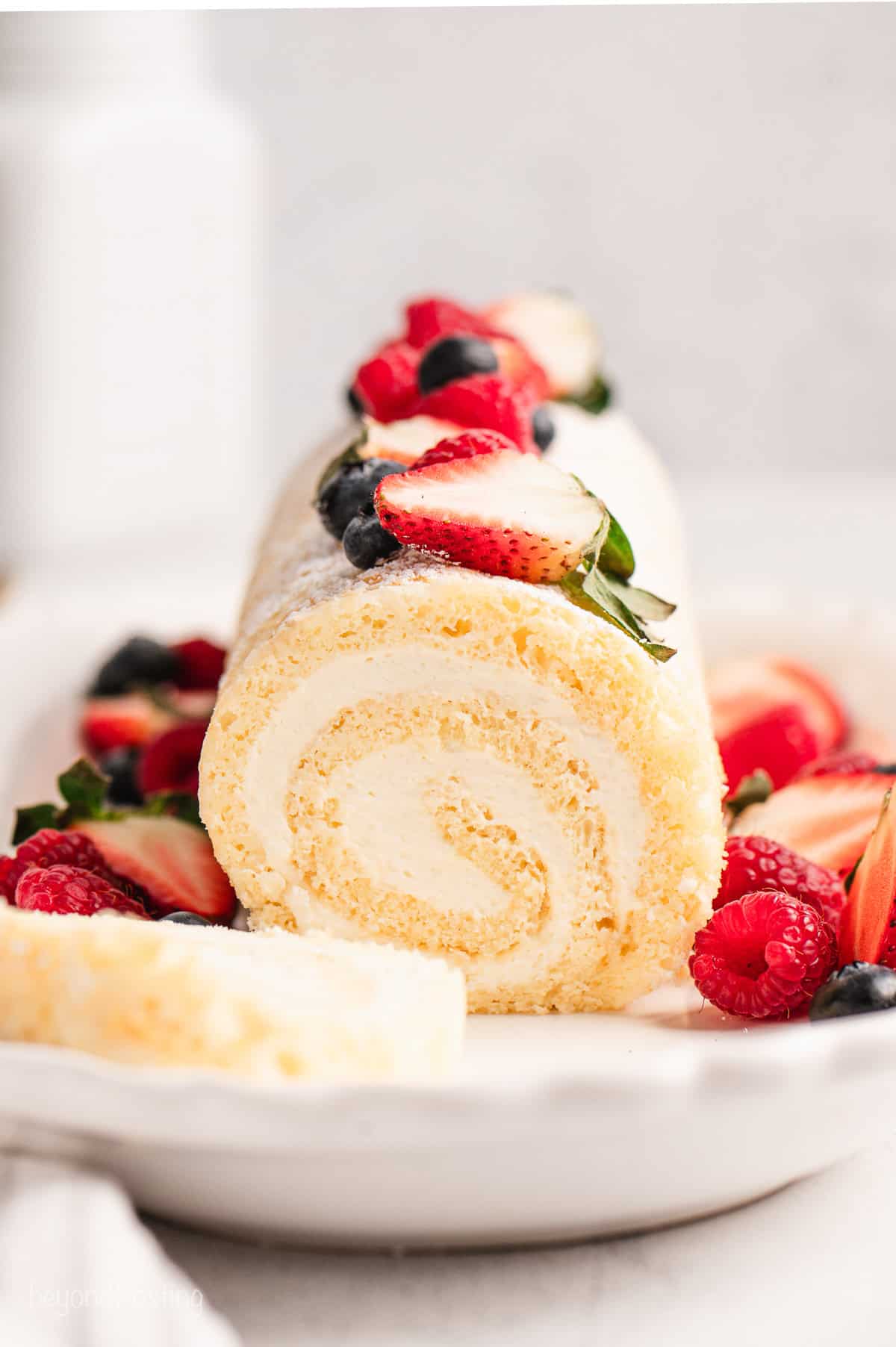 This easy vanilla cake roll is a soft and tender vanilla sponge cake swirled with tangy cream cheese frosting and dusted with powdered sugar. Front view of a vanilla cake roll topped with fresh mixed berries, with a slice cut from the end.