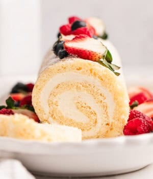 Front view of a vanilla cake roll topped with fresh mixed berries, with a slice cut from the end.