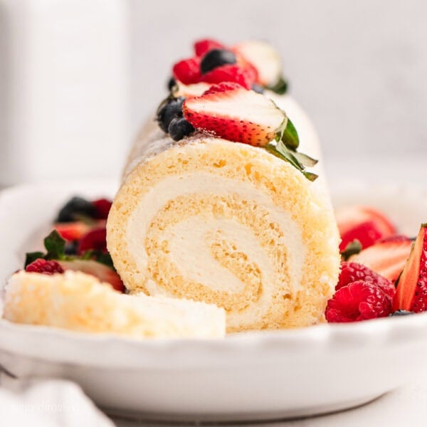 Front view of a vanilla cake roll topped with fresh mixed berries, with a slice cut from the end.