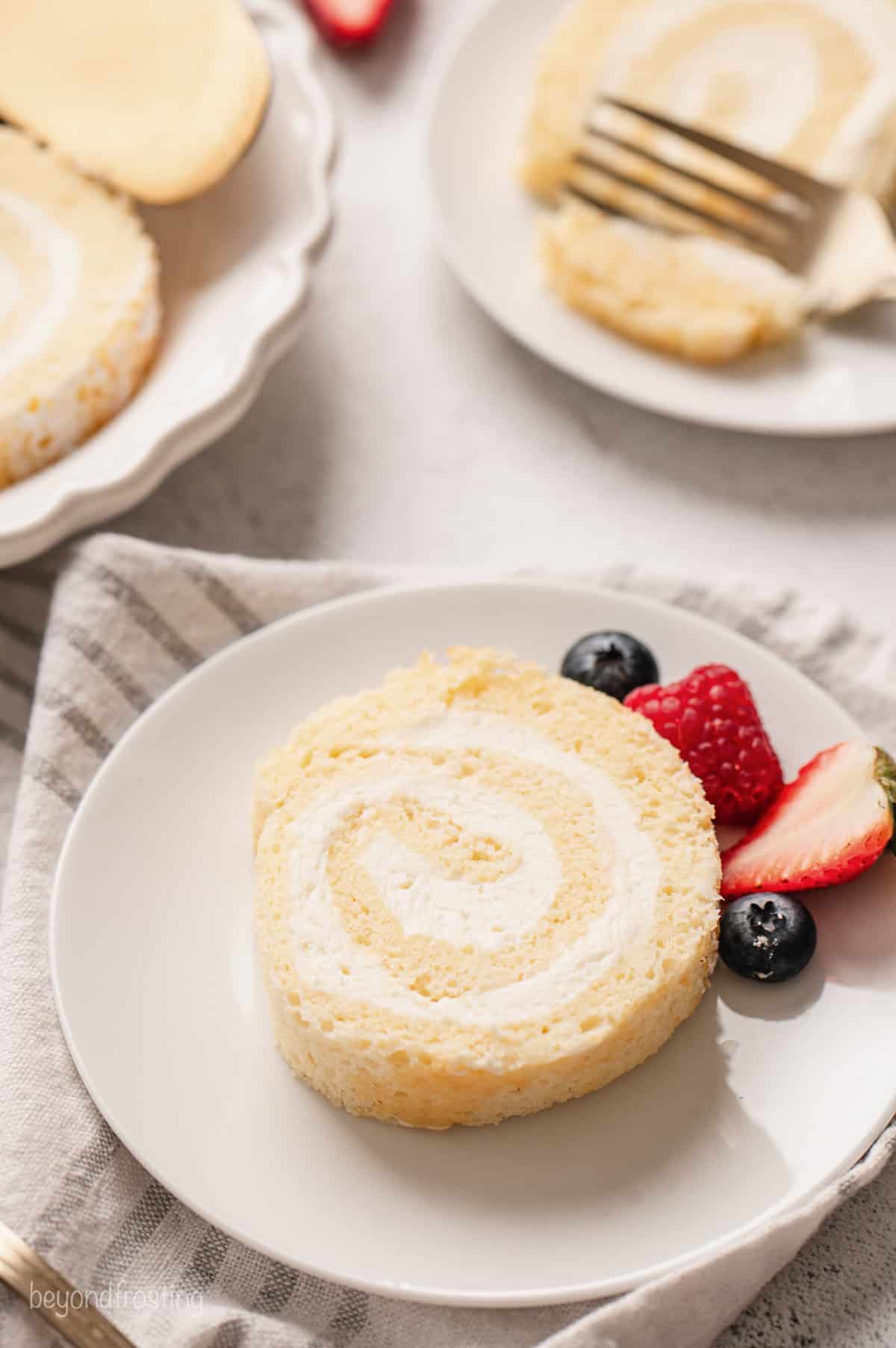 A slice of vanilla cake roll next to fresh mixed berries on a white plate.