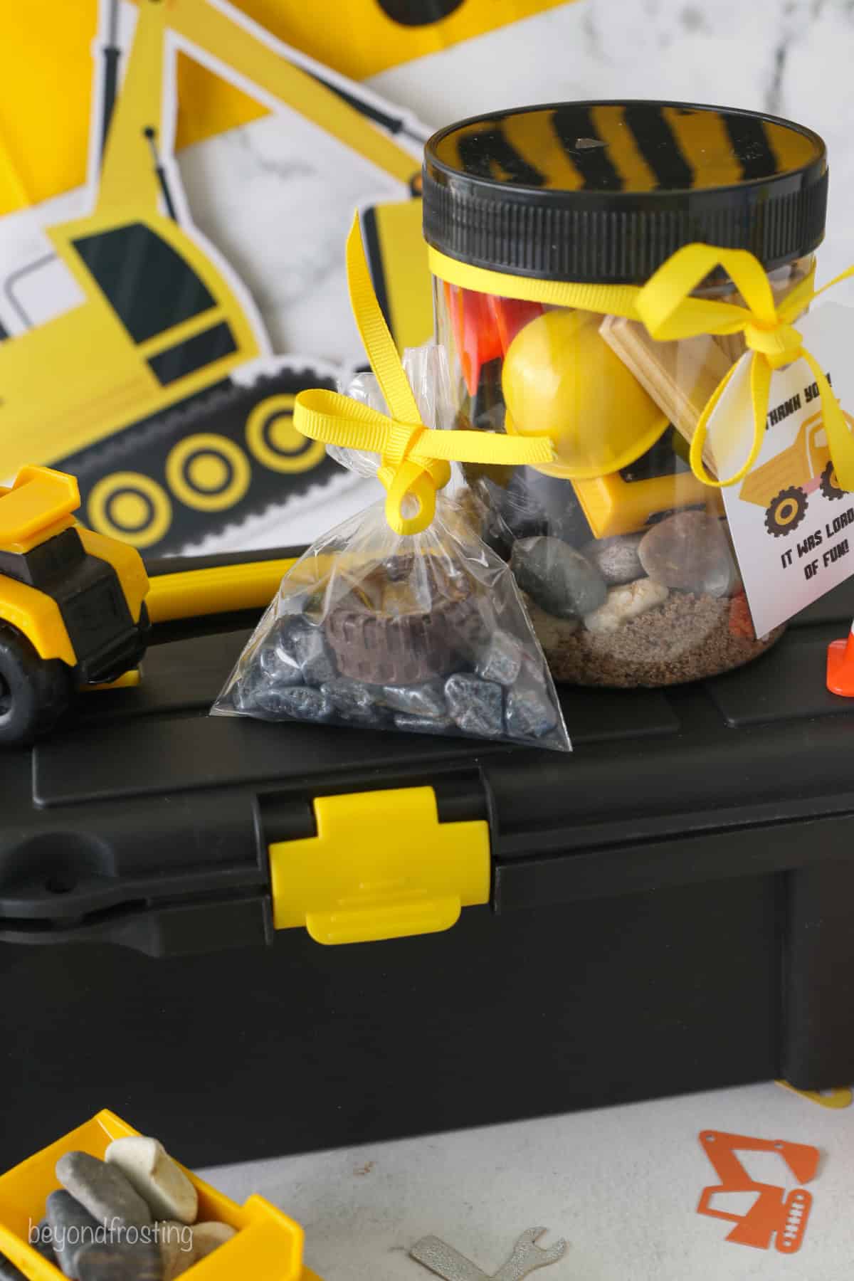 A small baggie of rocks and construction themed party favors next to a jar filled with kinetic sand and more party favors, on top of a toolbox.