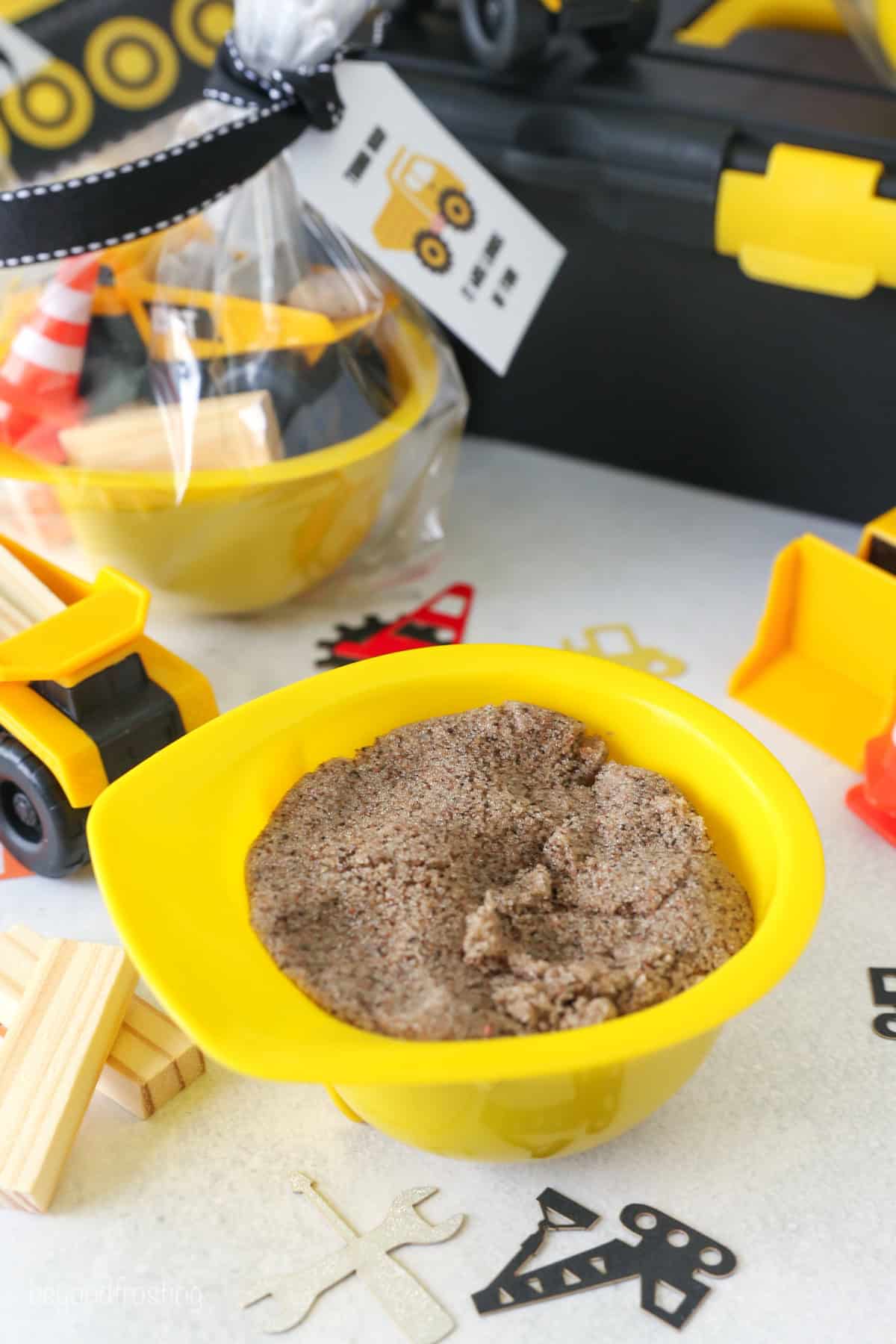 A yellow construction hat bowl filled with kinetic sand on a tabletop surrounded by party decorations.