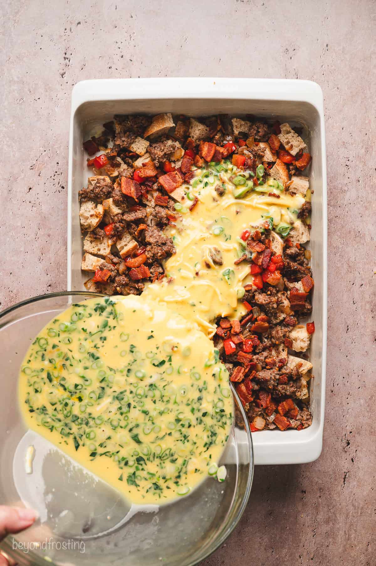 Pouring the egg mixture over the bread cubes and sausage in a baking dish.