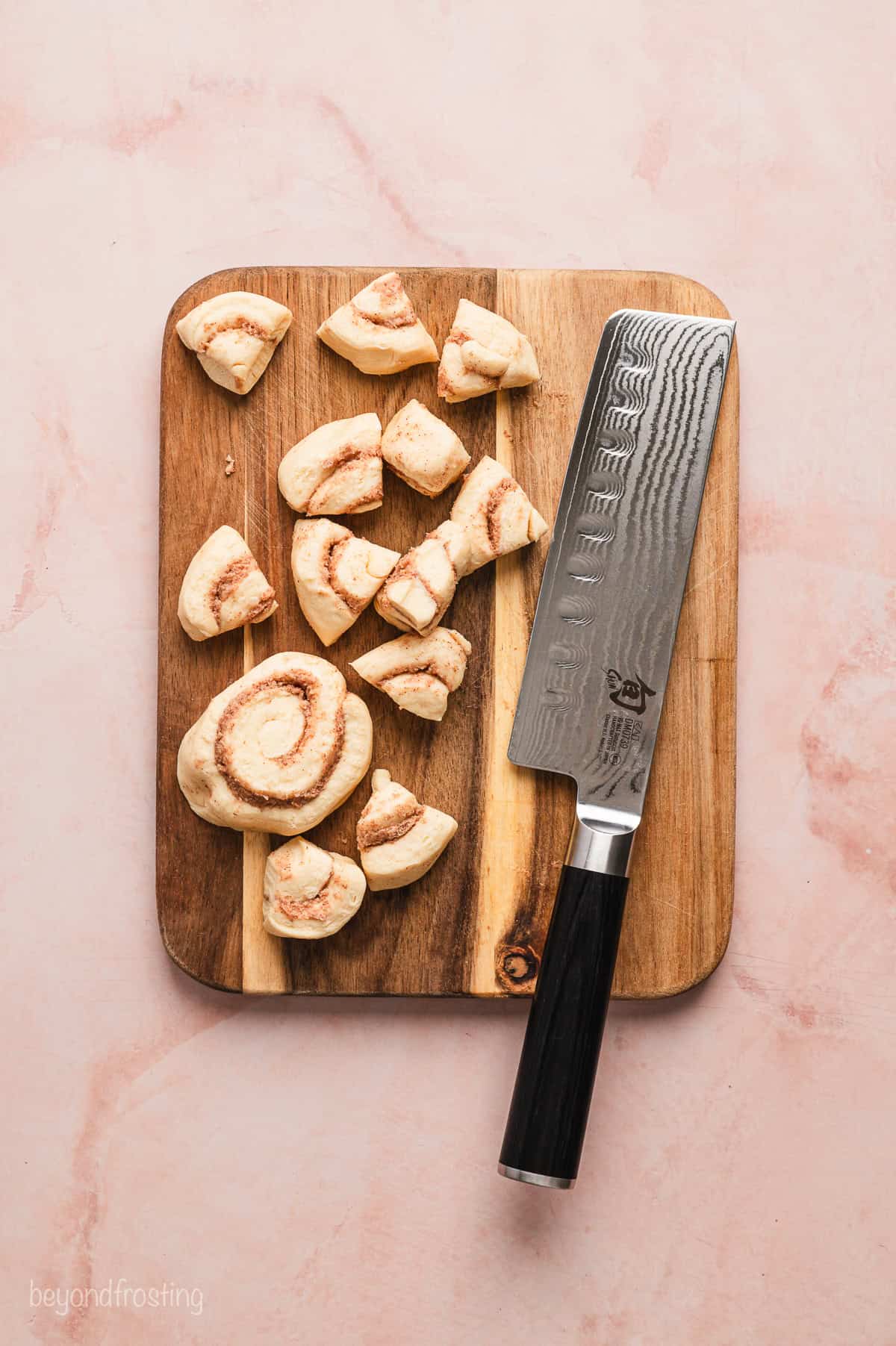Chopped refrigerated cinnamon rolls next to a large knife on a wooden cutting board.