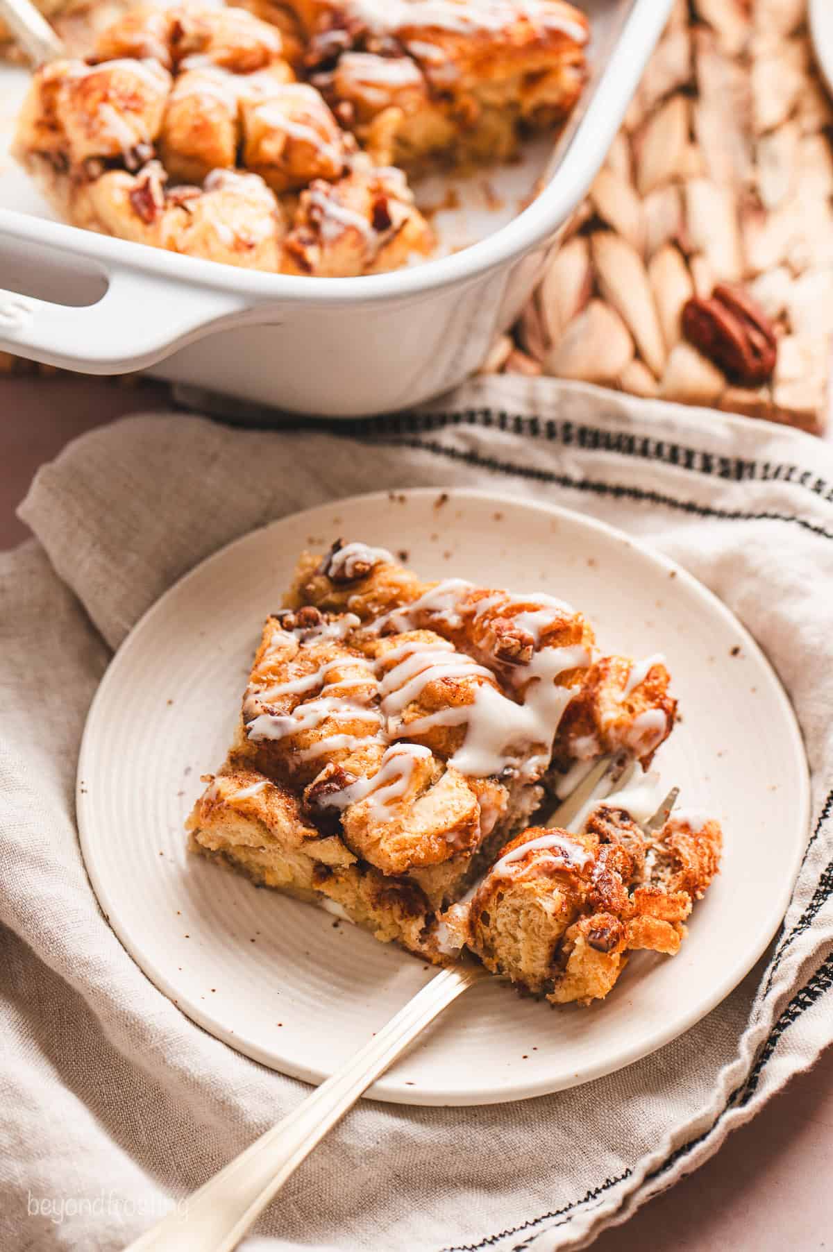 Overhead view of a slice of cinnamon roll casserole drizzled with icing on a plate, next to the rest of the casserole in a baking dish.