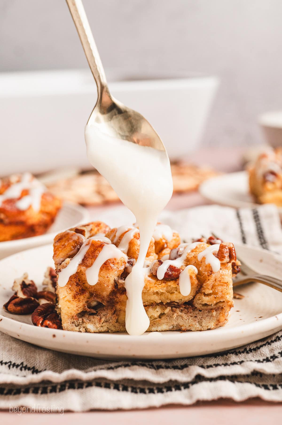 A spoon drizzling cream cheese icing over a serving of cinnamon roll casserole on a plate.