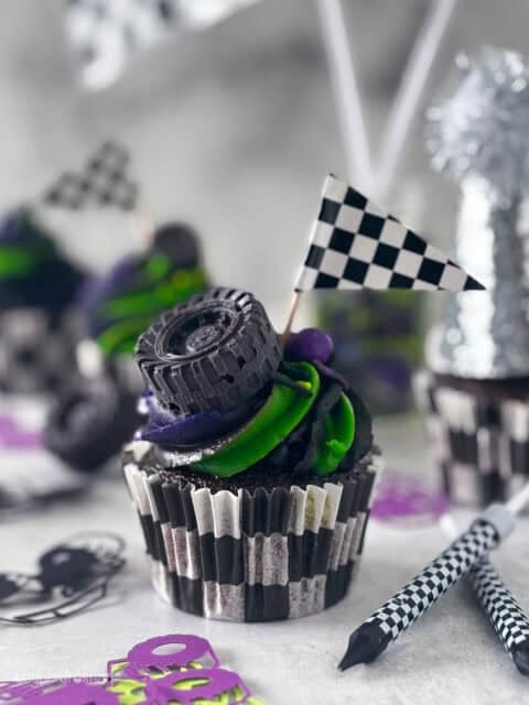 A chocolate cupcake with a swirled purple and green frosting decorated with a checkered flag and chocolate tires.