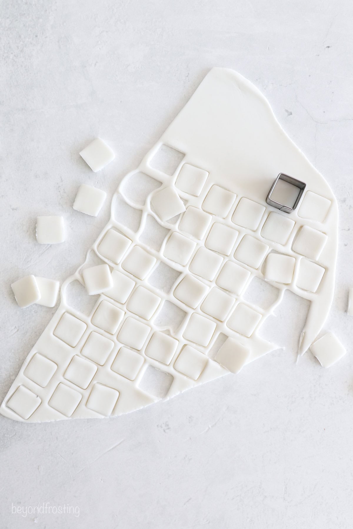 Overhead view of a small metal cutter cutting squares from a sheet of rolled out white fondant.