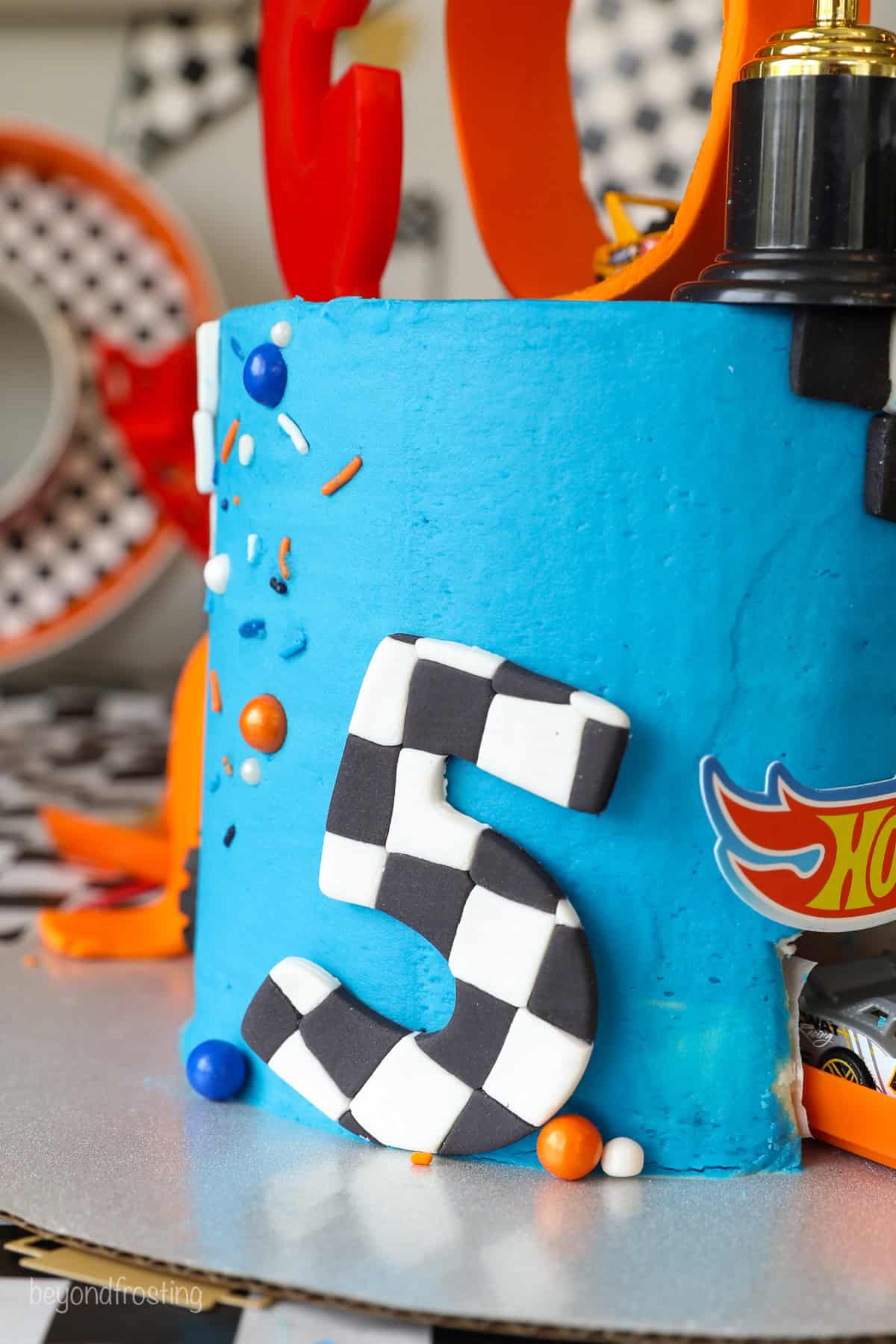 Side view of a Hot Wheels cake frosted with blue frosting, decorated with a number 5 made from checkered fondant.