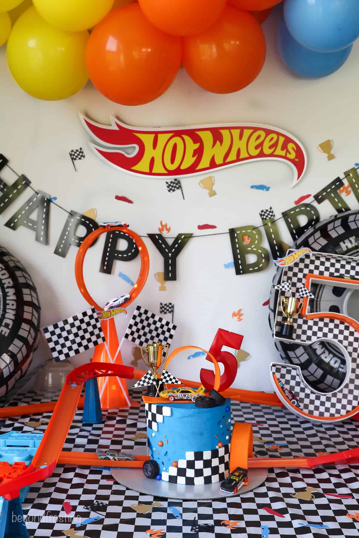 A Hot Wheels cake in the center of a table decorated with racing themed decorations with a Hot Wheels banner, happy birthday banner, and colorful balloons in the background.