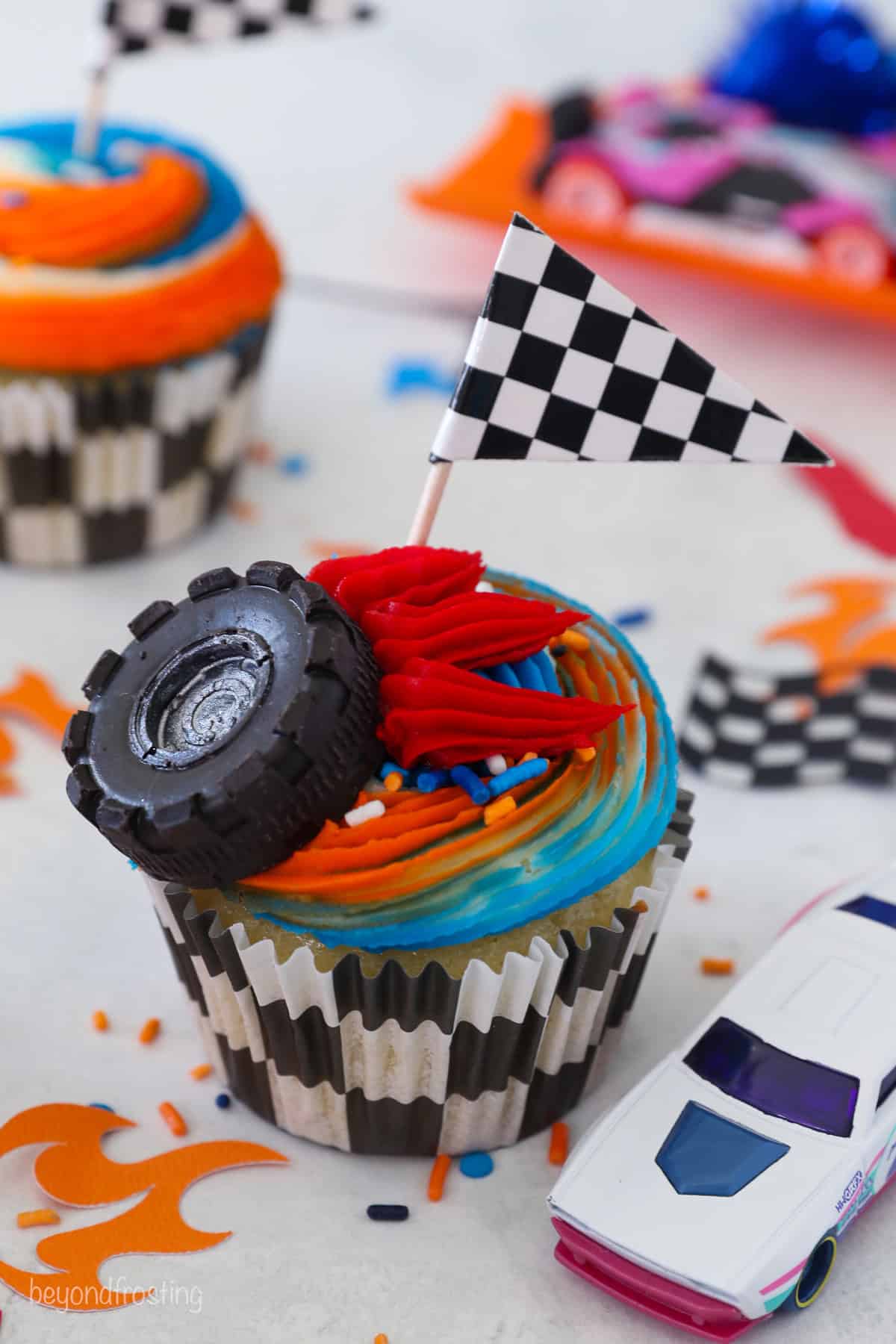 A Hot Wheels themed cupcake topped with colorful frosting, a chocolate tire, and a mini racing flag inside a cupcake liner with a checkered pattern.