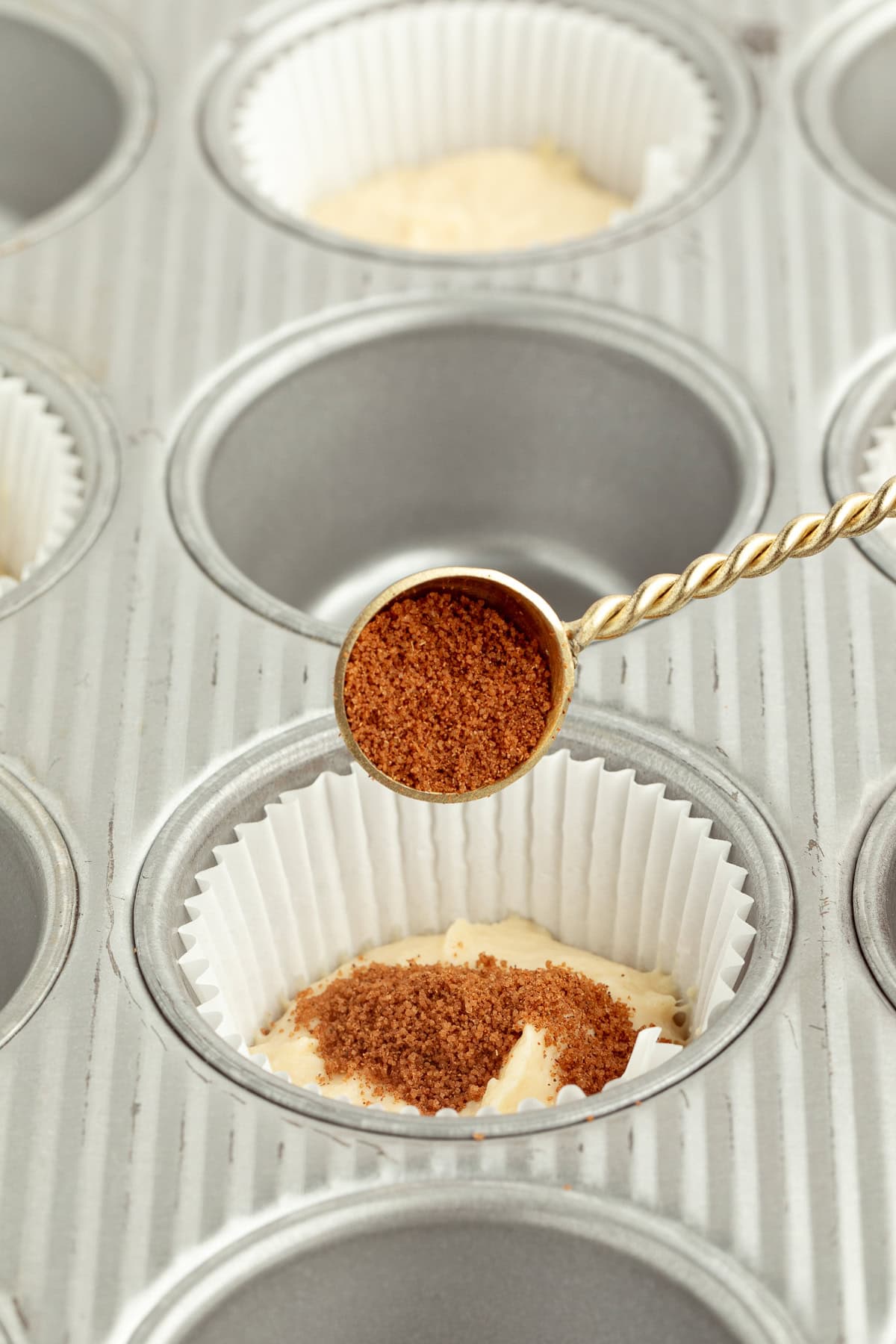 A teaspoon of cinnamon sugar filling being added on top of muffin batter inside a lined muffin pan.