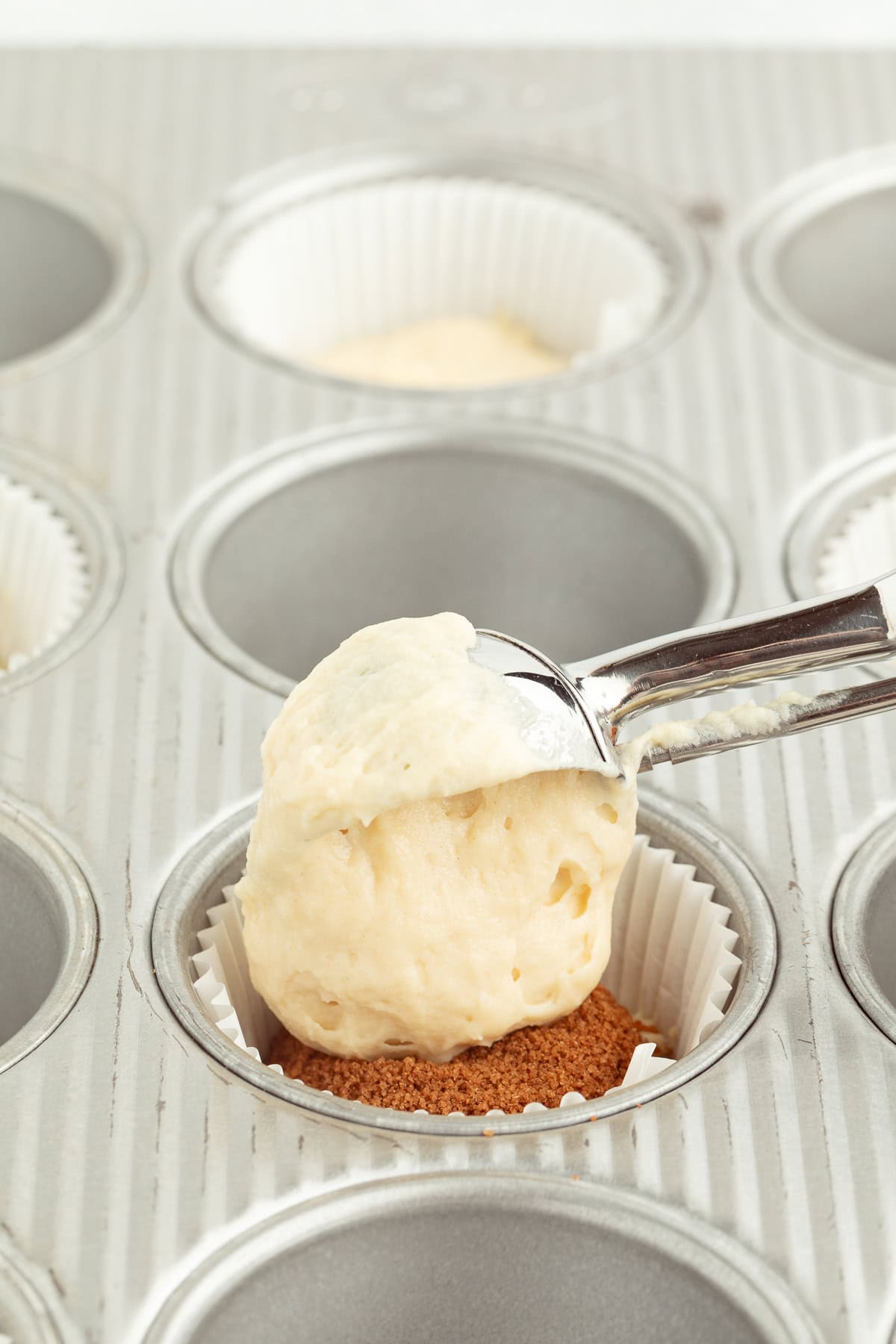An ice cream scoop dropping muffin batter on top of cinnamon filling in the well of a muffin pan.
