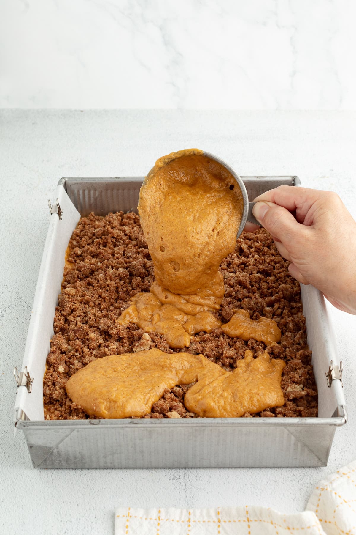 Pumpkin coffee cake batter being poured on top of the streusel layer inside a lined baking pan.