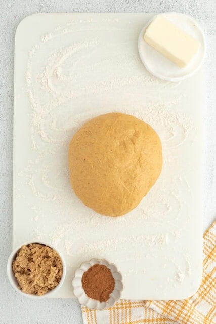 A ball of pumpkin cinnamon roll dough turned out on a floured cutting board next to small dishes of butter, brown sugar, and spices for the filling.