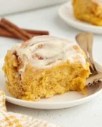 A pumpkin cinnamon roll with cream cheese icing on a plate next to a fork.