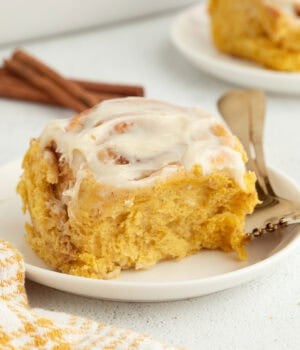 A pumpkin cinnamon roll with cream cheese icing on a plate next to a fork.