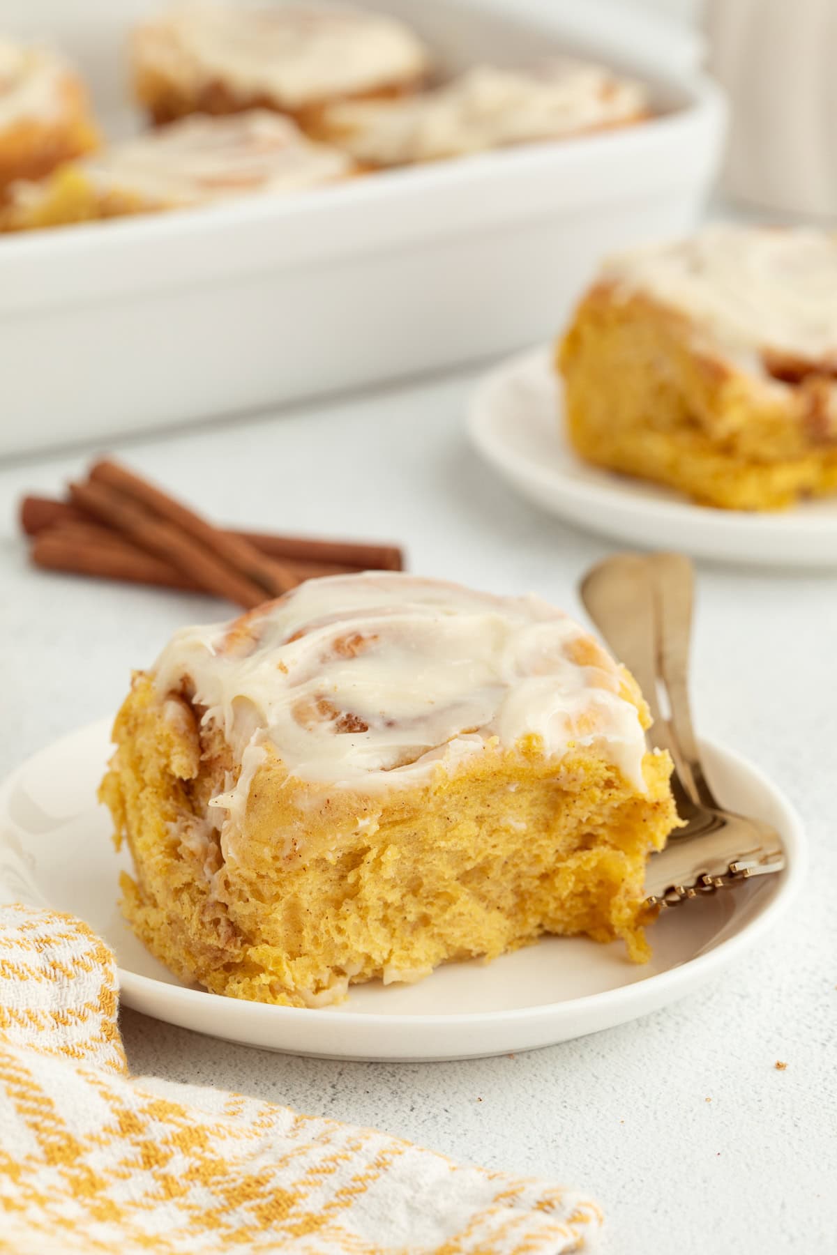 These soft, fluffy pumpkin cinnamon rolls made with real pumpkin purée, warm spices, and cream cheese frosting are perfect for fall mornings. A pumpkin cinnamon roll with cream cheese icing on a plate next to a fork, with a second cinnamon roll in the background.