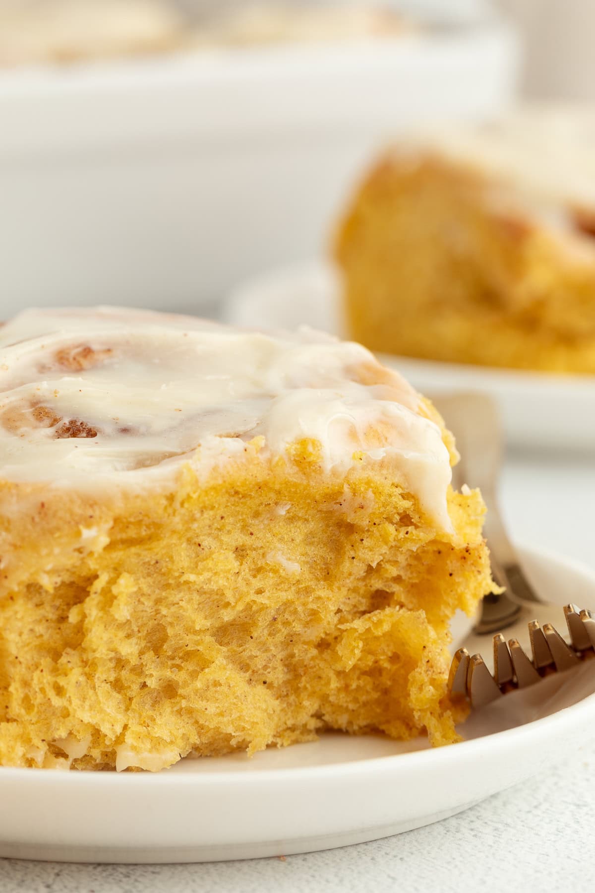 Close up of a pumpkin cinnamon roll with cream cheese icing on a plate next to a fork, with a second cinnamon roll in the background.