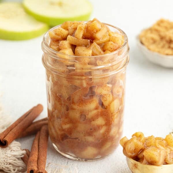 Stovetop apple pie filling in a glass mason jar next to cinnamon sticks and sliced apples.