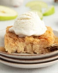 A slice of homemade apple bread pudding topped with ice cream on a plate next to a fork.