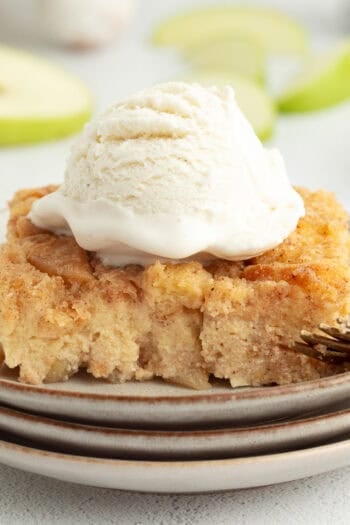 A slice of homemade apple bread pudding topped with ice cream on a plate next to a fork.