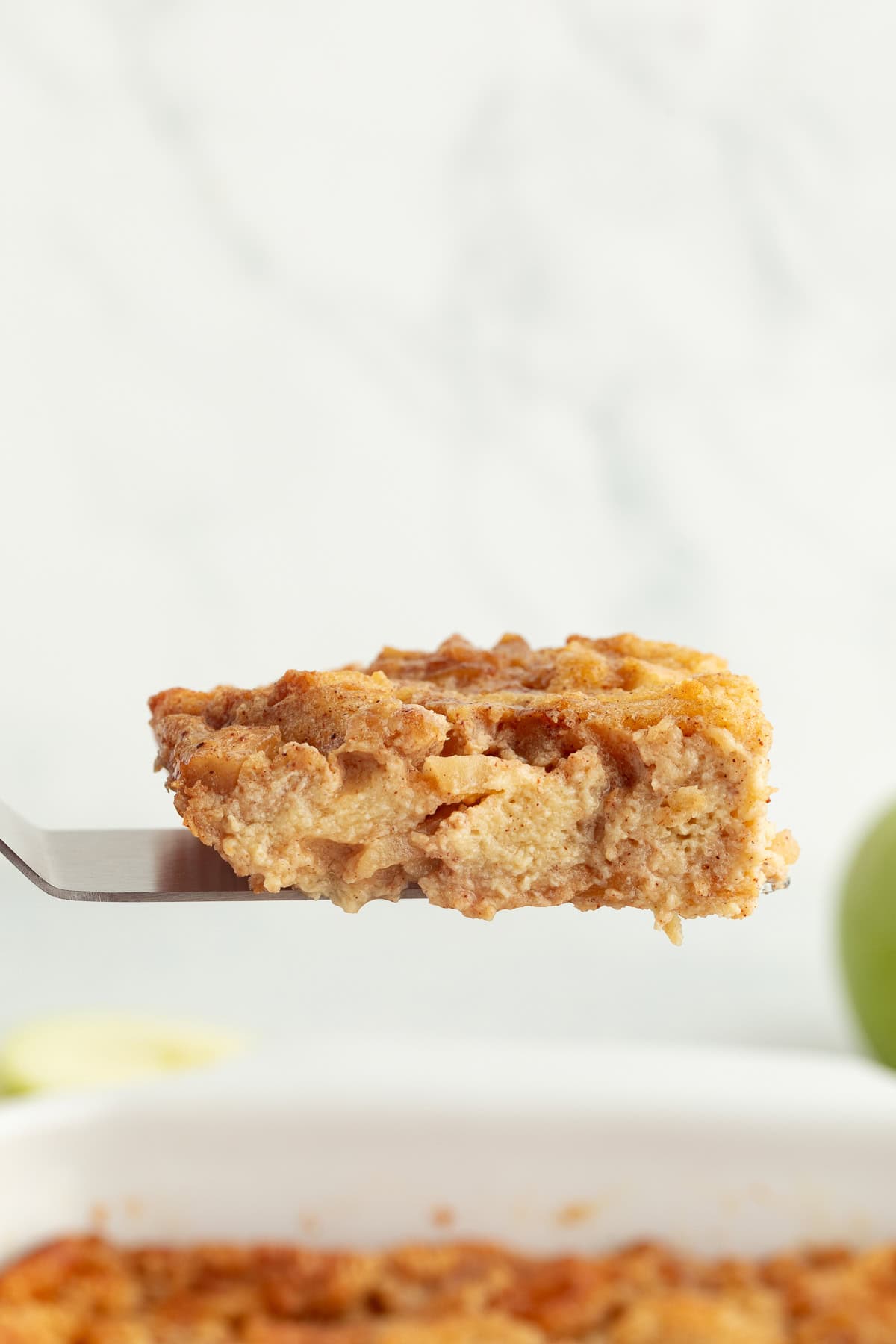 A slice of apple pie bread pudding being lifted from a baking dish with a spatula.