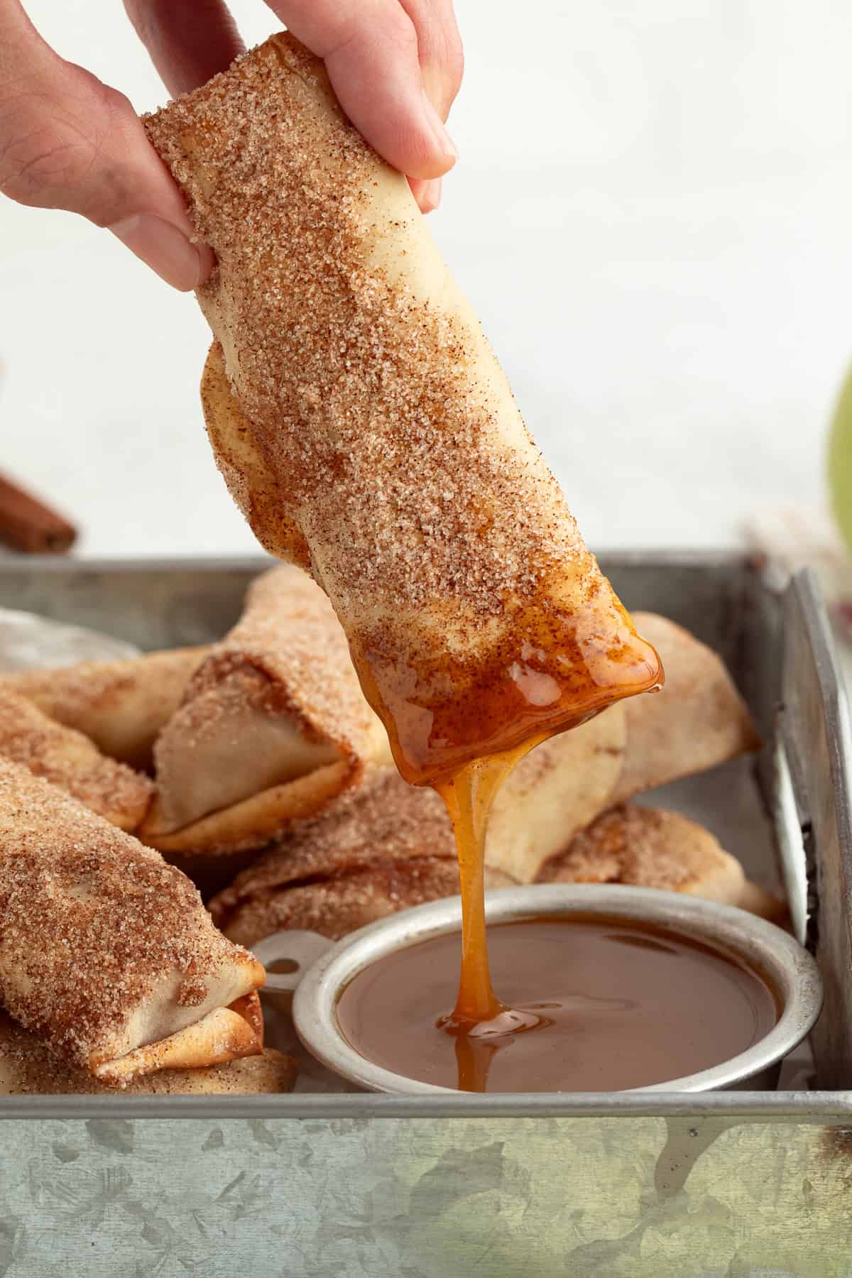 These apple pie egg rolls are golden, crispy egg roll wrappers bursting with homemade apple pie filling. Make them in the oven or air fryer! A hand dipping an apple pie egg roll into a small ramekin of caramel sauce, surrounded by more egg rolls on a tray.