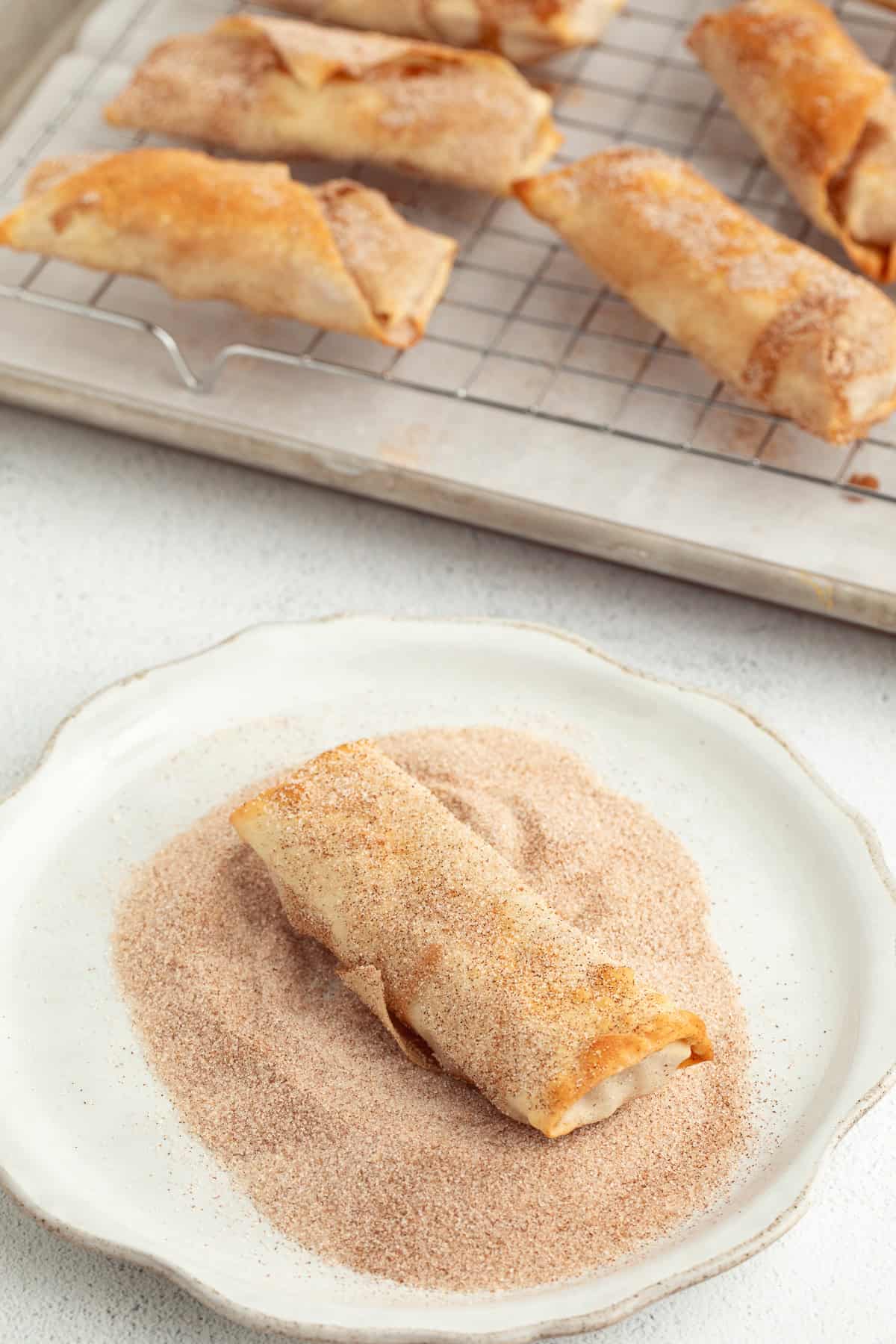 These apple pie egg rolls are golden, crispy egg roll wrappers bursting with homemade apple pie filling. Make them in the oven or air fryer! An apple pie egg roll in a shallow bowl of cinnamon sugar, with more egg rolls on a wire rack in the background.