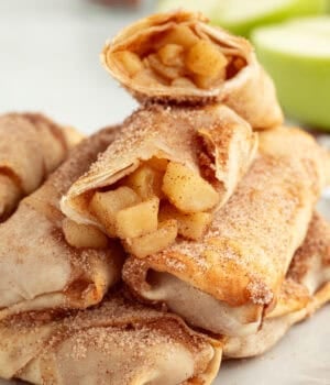 Apple pie egg rolls stacked on a plate, with the top egg roll broken in half to reveal the juicy apple pie filling inside.