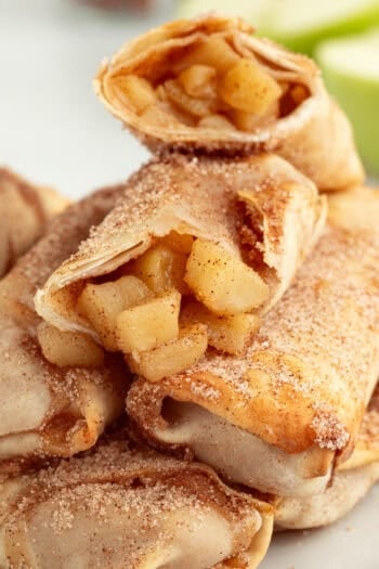 Apple pie egg rolls stacked on a plate, with the top egg roll broken in half to reveal the juicy apple pie filling inside.