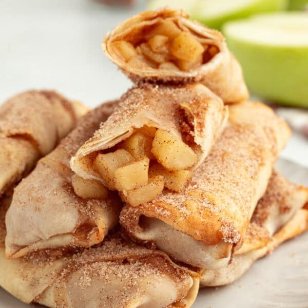 Apple pie egg rolls stacked on a plate, with the top egg roll broken in half to reveal the juicy apple pie filling inside.
