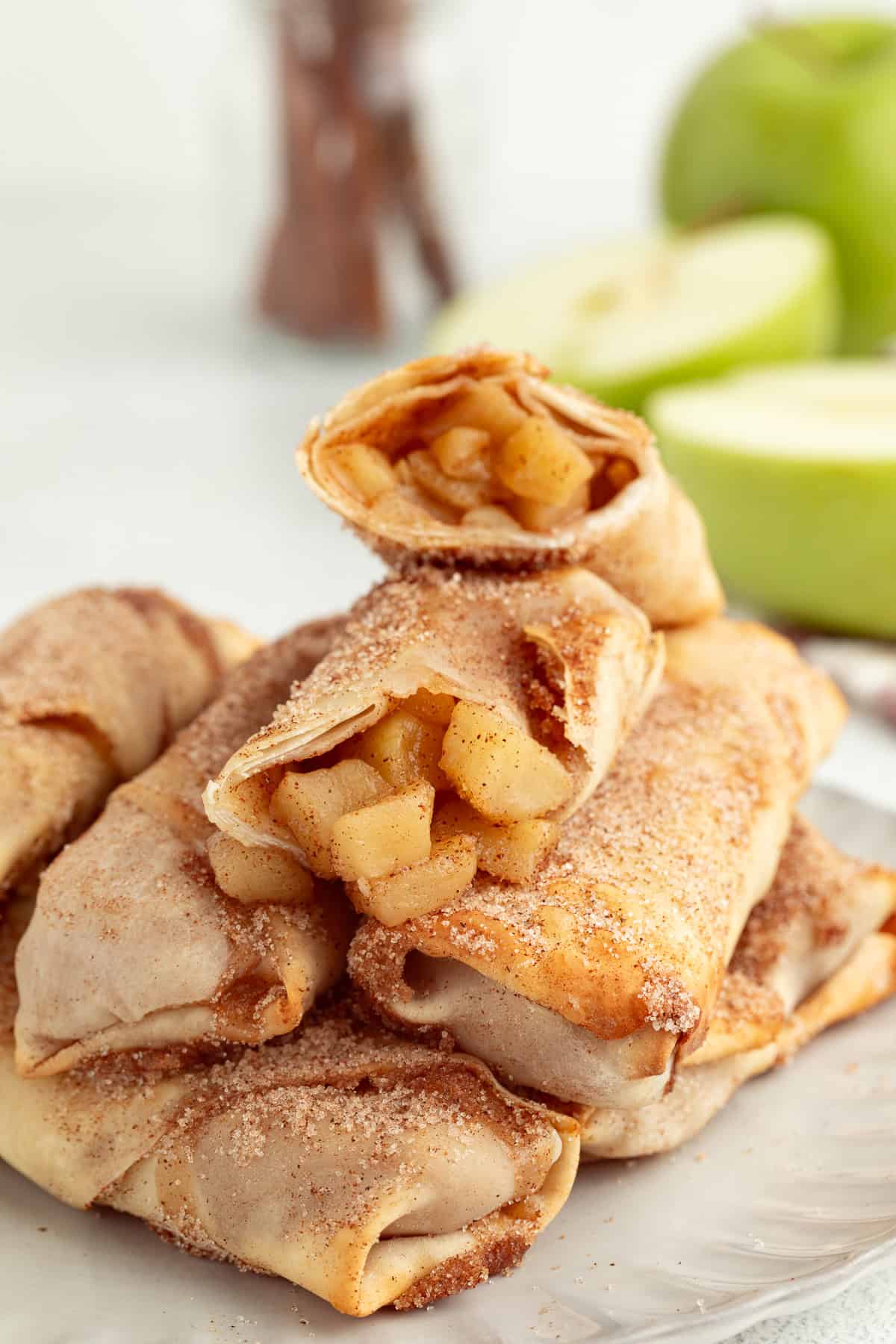 These apple pie egg rolls are golden, crispy egg roll wrappers bursting with homemade apple pie filling. Make them in the oven or air fryer! Apple pie egg rolls stacked on a plate, with the top egg roll broken in half to reveal the juicy apple pie filling inside.
