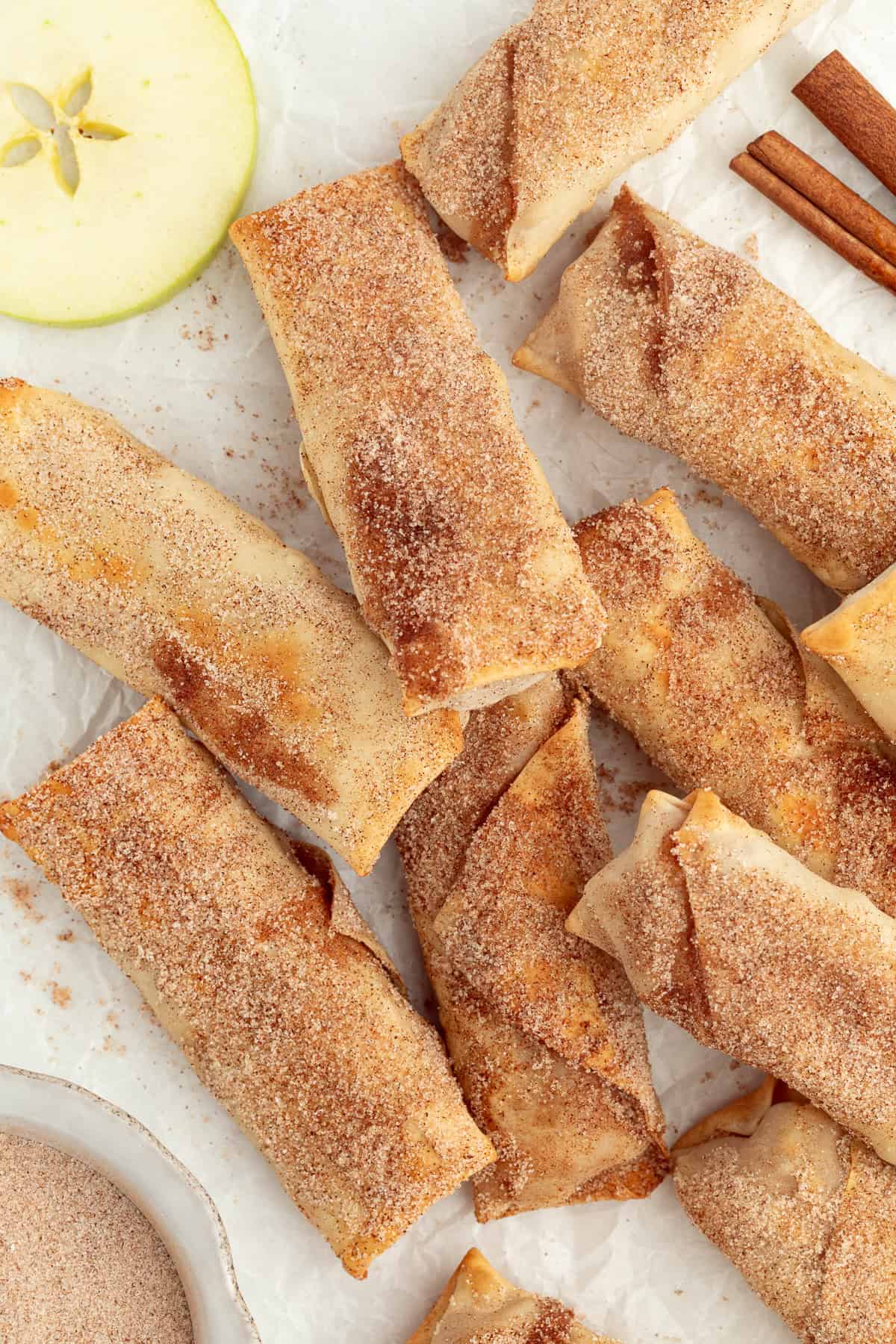 These apple pie egg rolls are golden, crispy egg roll wrappers bursting with homemade apple pie filling. Make them in the oven or air fryer! Overhead view of assorted apple pie egg rolls coated with cinnamon sugar.