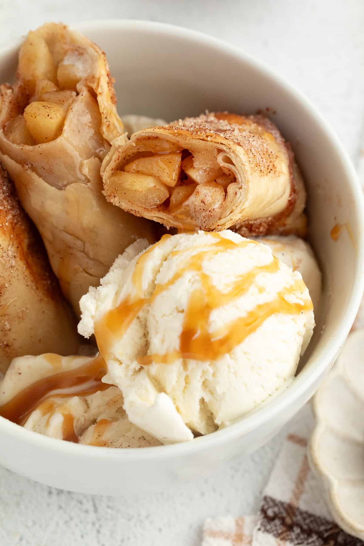 These apple pie egg rolls are golden, crispy egg roll wrappers bursting with homemade apple pie filling. Make them in the oven or air fryer! Apple pie egg rolls tucked into a bowl next to scoops of vanilla ice cream, drizzled with caramel sauce.