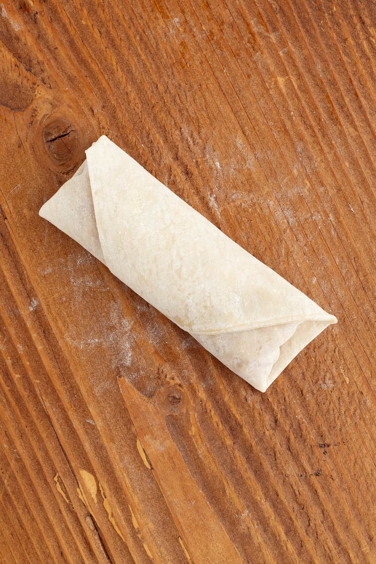 These apple pie egg rolls are golden, crispy egg roll wrappers bursting with homemade apple pie filling. Make them in the oven or air fryer! Close up of a wrapped apple pie egg roll on a wooden surface.