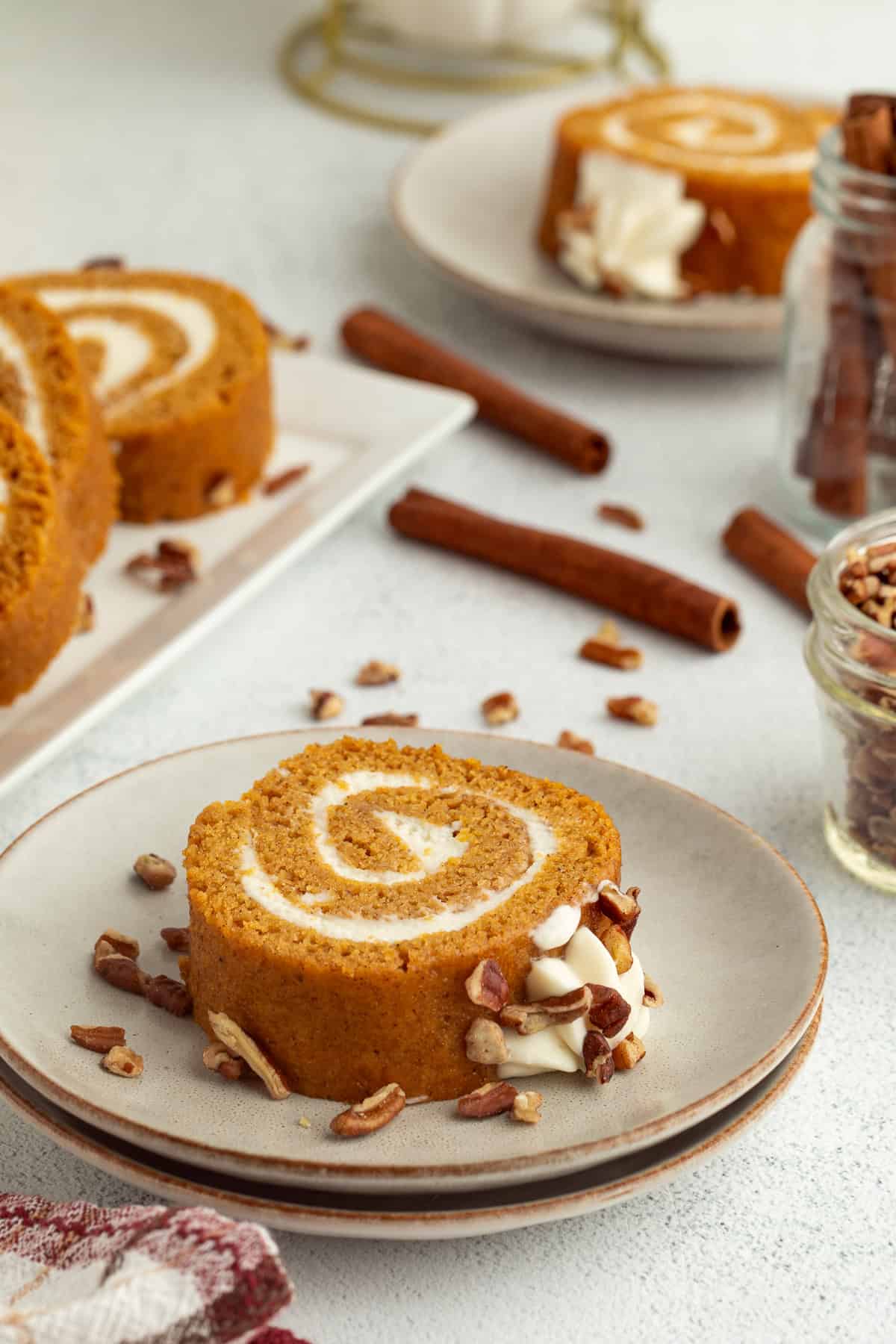 A slice of pumpkin cake roll garnished with whipped cream and chopped pecans on a plate, with more cake slices in the background.