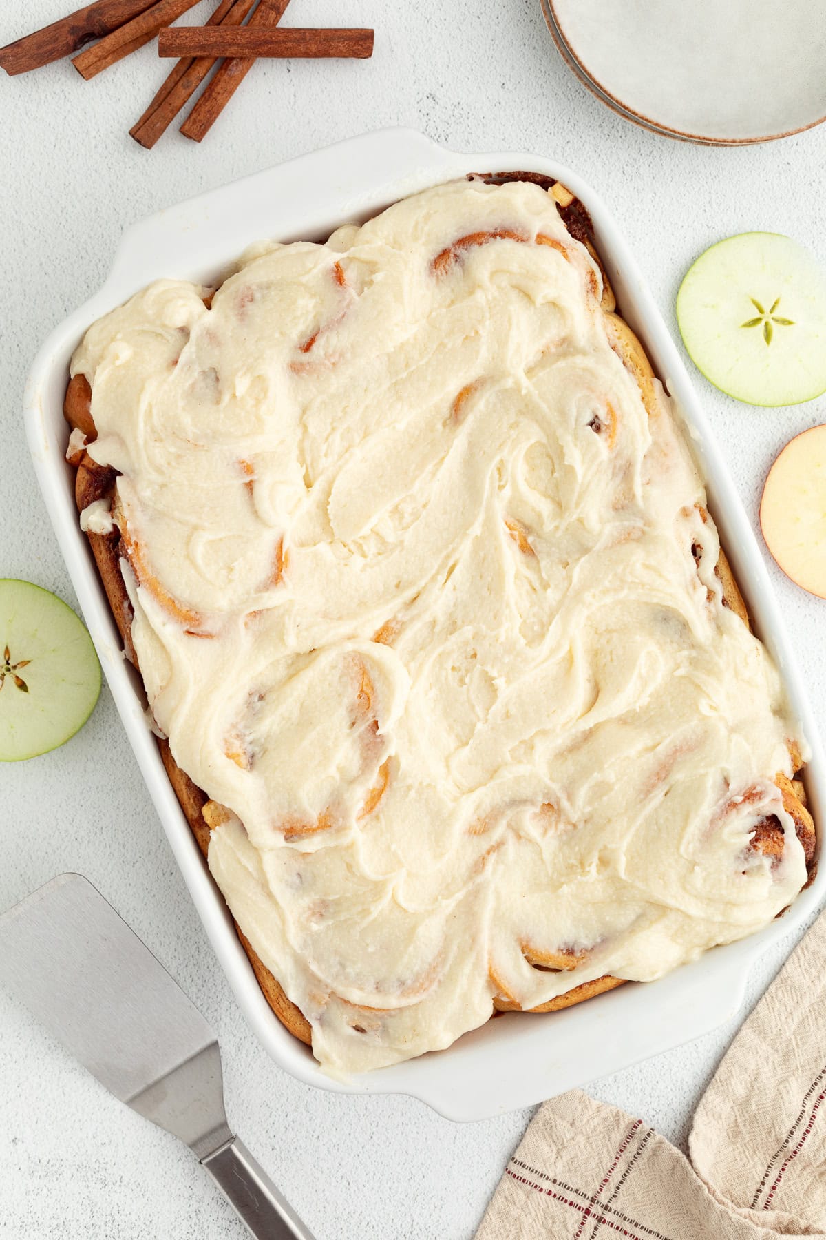 Homemade apple cinnamon rolls are golden-baked with warm cinnamon apple filling and topped with creamy brown butter cream cheese frosting. Overhead view of apple cinnamon rolls topped with brown butter cream cheese frosting in a baking dish.