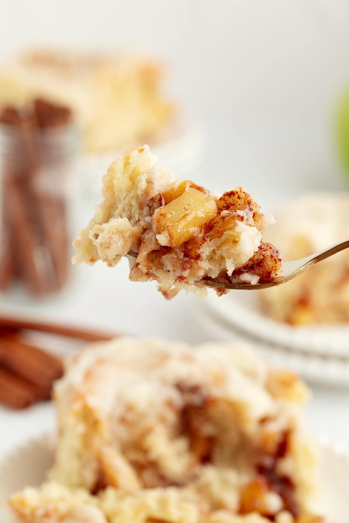 Homemade apple cinnamon rolls are golden-baked with warm cinnamon apple filling and topped with creamy brown butter cream cheese frosting. A forkful of apple cinnamon roll held over the rest of the cinnamon roll on a plate.