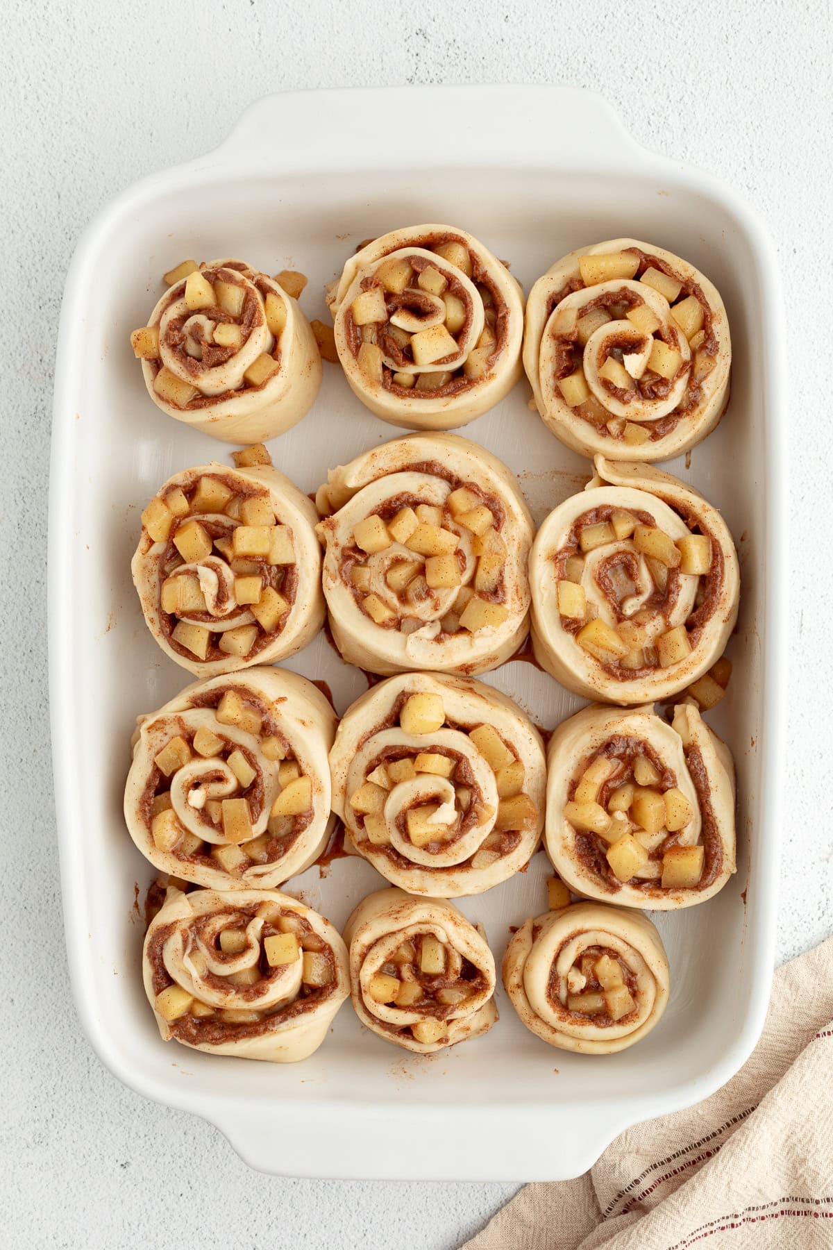 Homemade apple cinnamon rolls are golden-baked with warm cinnamon apple filling and topped with creamy brown butter cream cheese frosting. Overhead view of unbaked apple cinnamon rolls in a baking dish.