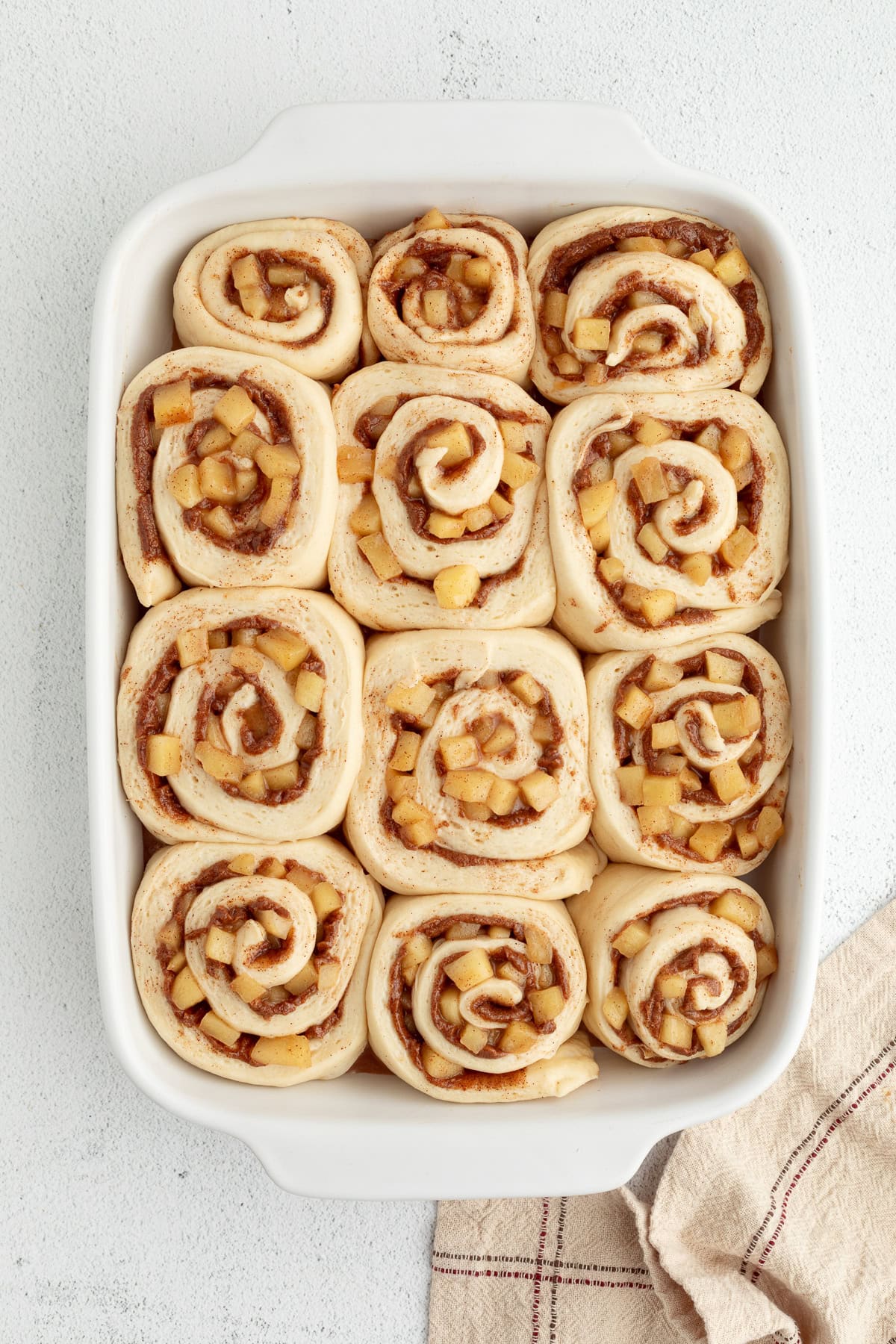 Homemade apple cinnamon rolls are golden-baked with warm cinnamon apple filling and topped with creamy brown butter cream cheese frosting. Overhead view of unbaked apple cinnamon rolls in a baking dish after rising.