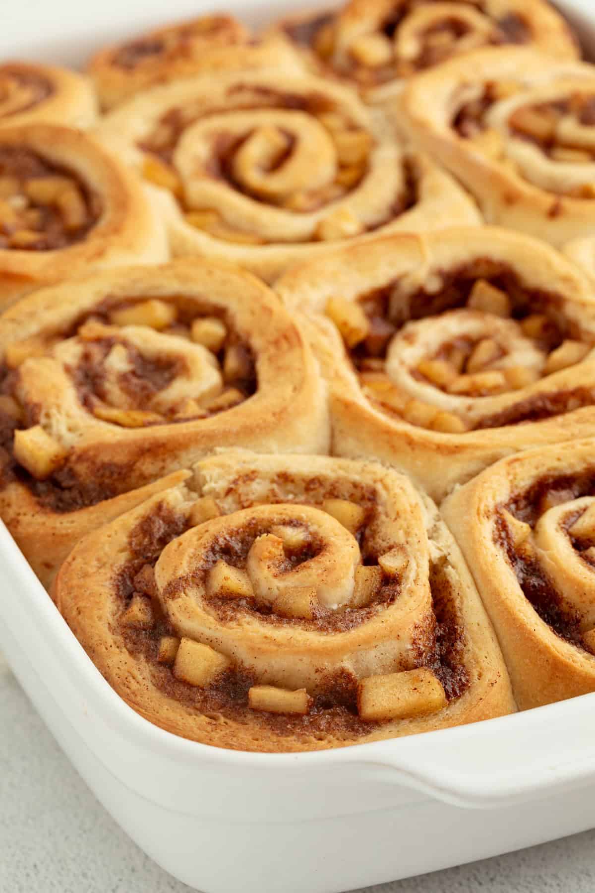 Homemade apple cinnamon rolls are golden-baked with warm cinnamon apple filling and topped with creamy brown butter cream cheese frosting. Close up of apple cinnamon rolls in a baking dish.
