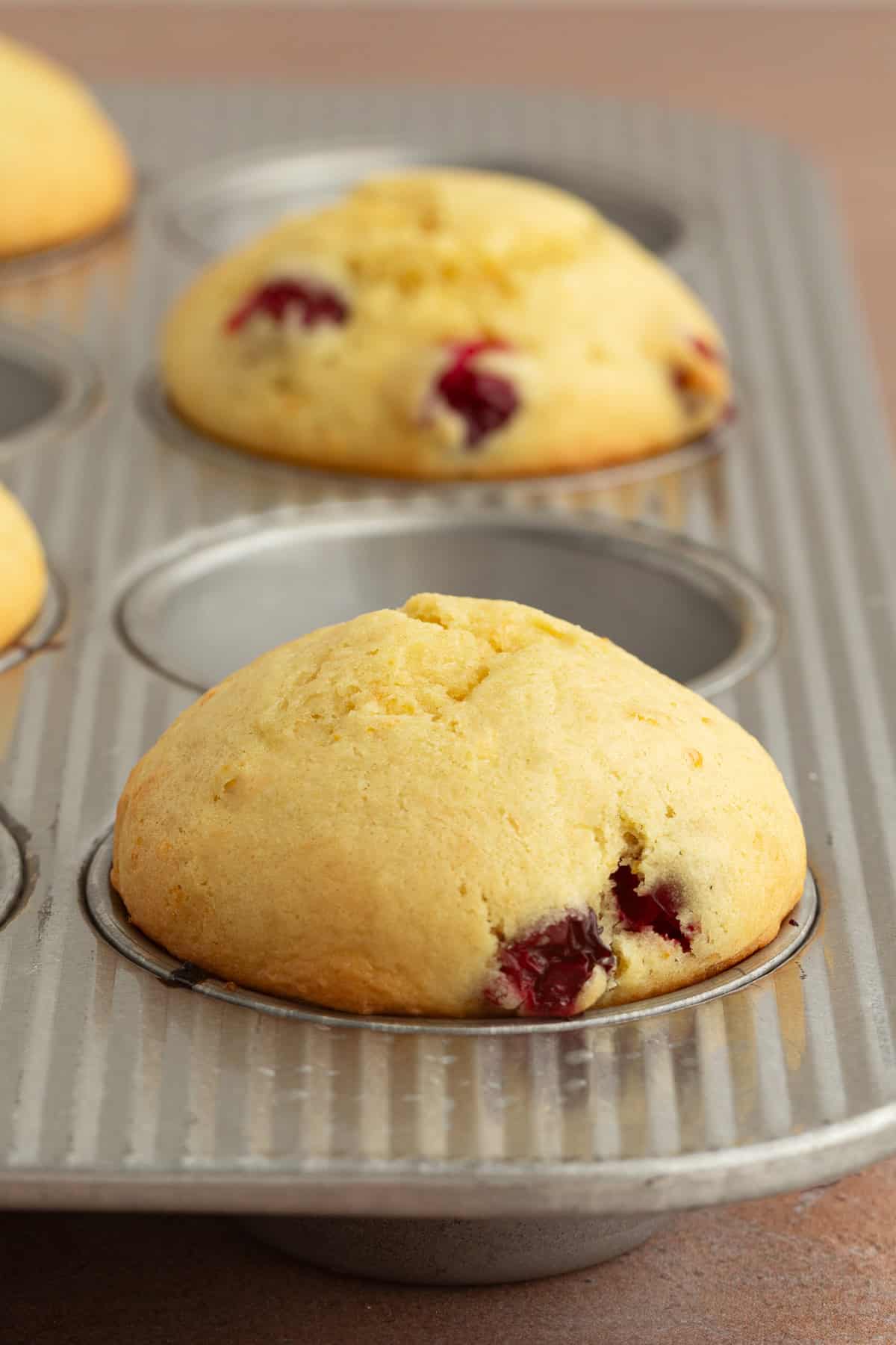 These moist cranberry orange muffins are full of real orange juice, fresh zest, and sweet-tart cranberries. So easy to make and big on flavor! Close up of baked orange cranberry muffins in a muffin tin, with an empty well between each muffin to allow for spreading.