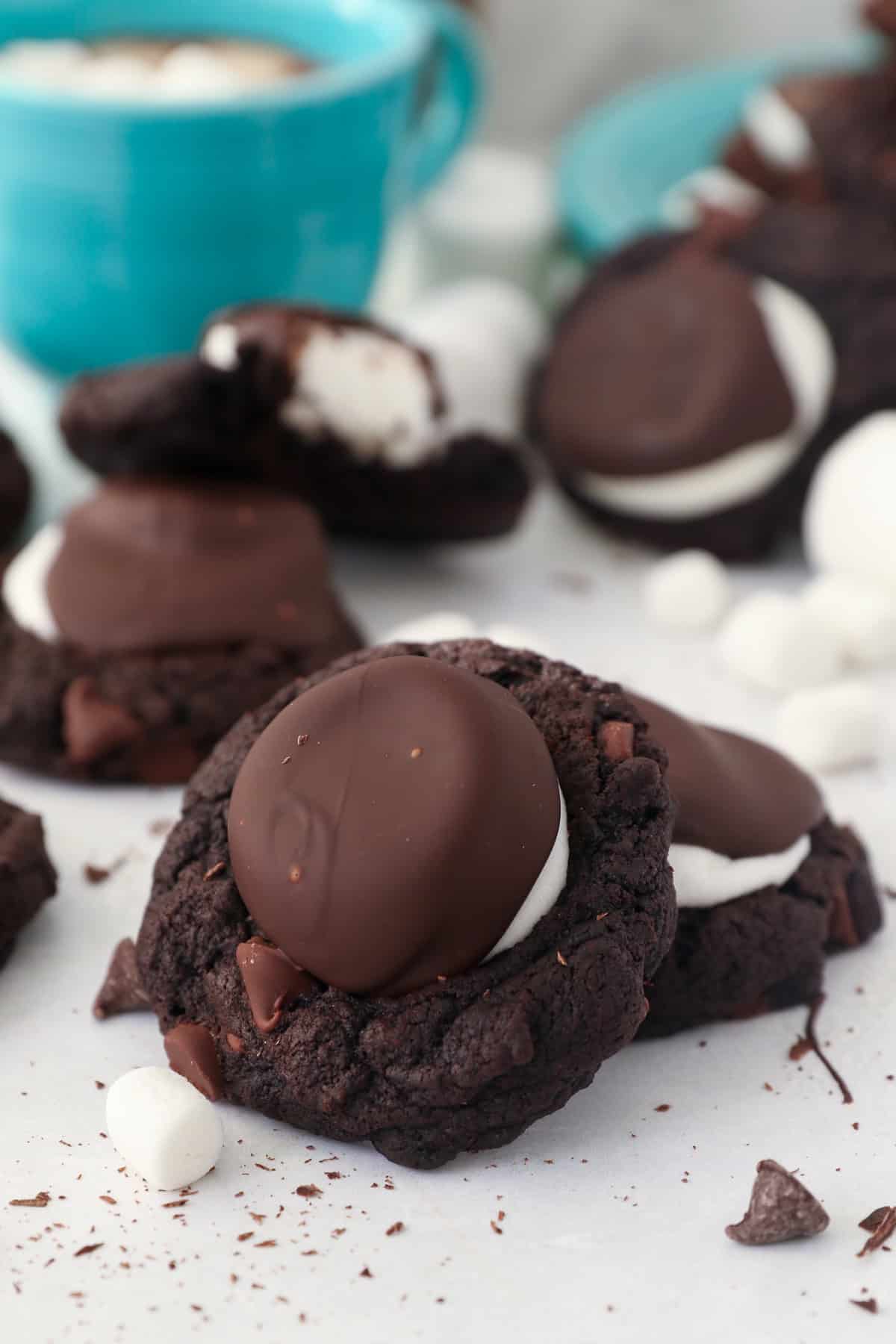 A close up of a chocolate covered marshmallow hot chocolate cookie leaned up again another cookie with more cookies in the background and a teal mug of hot chocolate