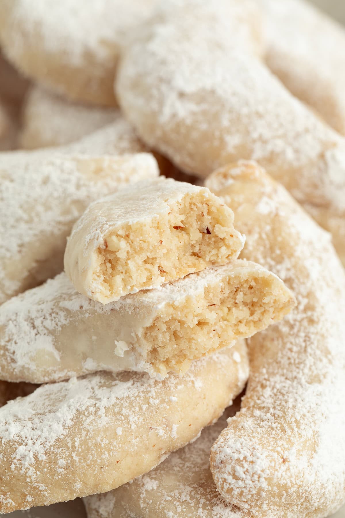 These classic almond crescent cookies have a buttery, crumbly shortbread texture and a sweet powdered sugar coating. No chilling needed! Close up of almond crescent cookies piled on a plate, with one cookie broken in half to show the crumbly interior.